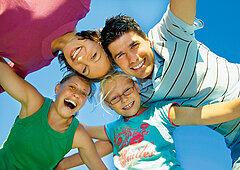 Familie: Eine Frau und ein Mann sind lachend mit ihren zwei Töchtern abgebildet. Im Hintergrund ist nur ein blauer Himmel zu sehen.