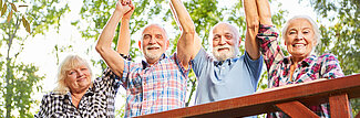 Fröhliche Senioren halten sich an den Händen und jubeln, im Hintergrund ist Natur zu sehen
